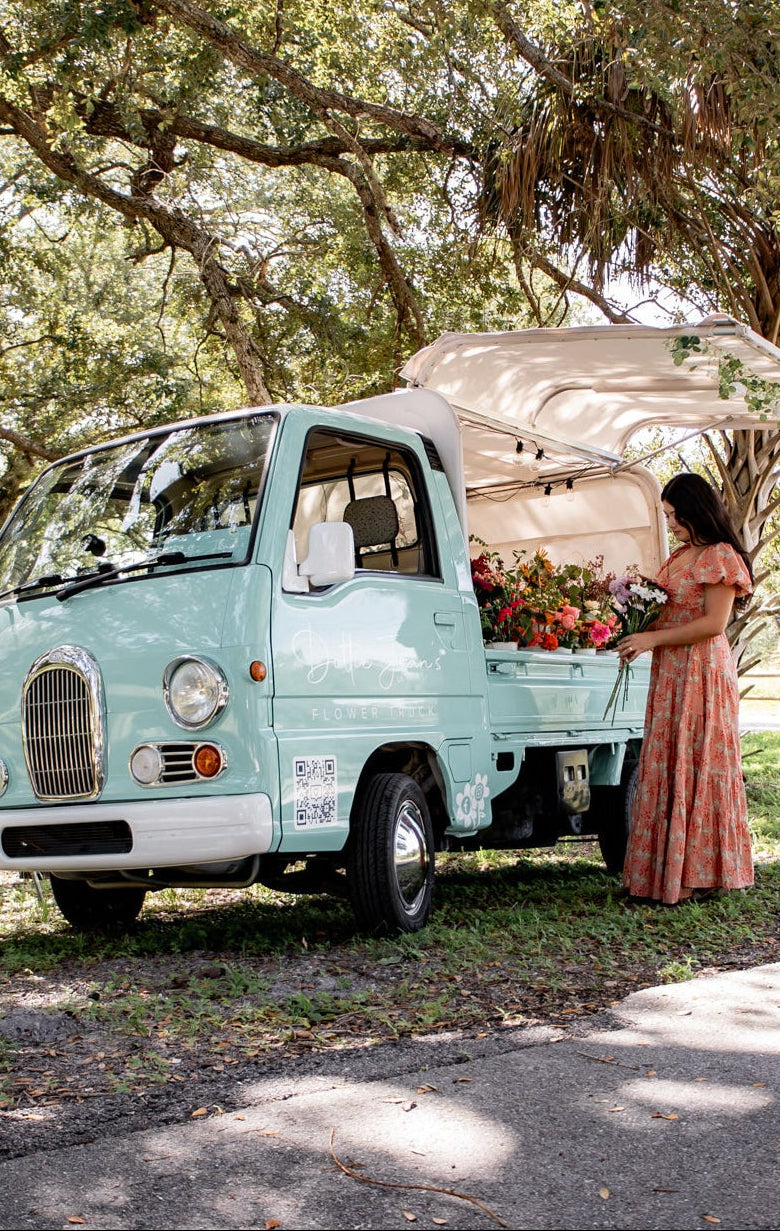 Vintage-style food truck under a tree with a woman in a long dress standing next to it.